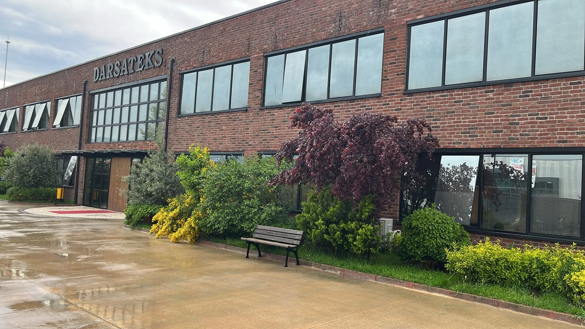 Front of the red-brick Darsateks factory in Erbaa, with flowering shrubs, a bench and a rain-wet forecourt
