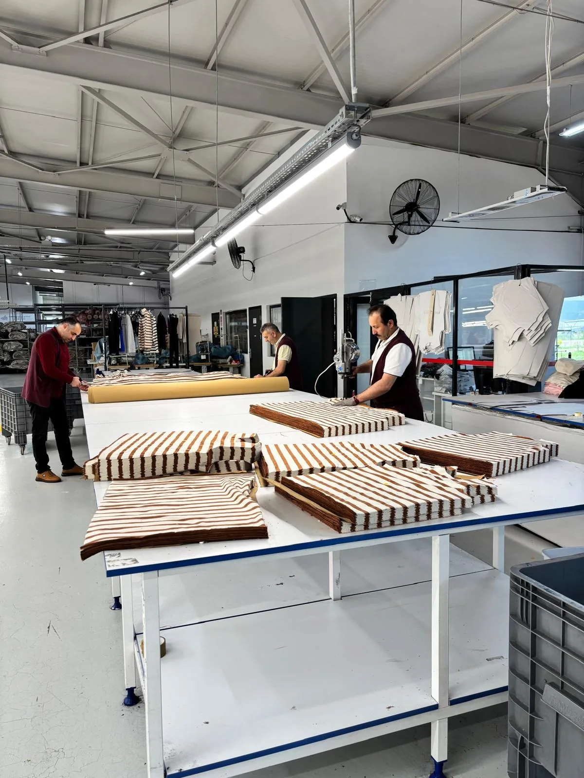 Stacks of striped knit panels on a finishing table in the Erbaa factory, with staff working in the hall behind